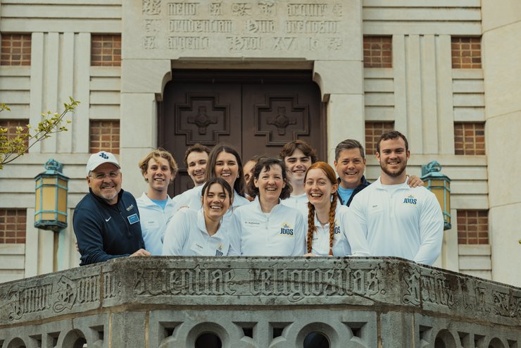 A group of nine people wearing white shirts with logos smile and pose together on a stone balcony in front of a large wooden.