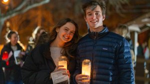 A young woman and man smile while holding lit candles outdoors at night with blurred people and trees in the background.