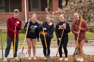 Five young people stand outside holding gardening tools and smiling in front of a chain-link fence and a brick building.