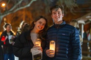 A young woman and man smile while holding lit candles outdoors at night with blurred people and trees in the background.