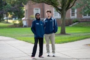 Two young adults wearing blue jackets stand smiling on a paved path with grass and a tree behind them.