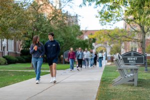 Two young people walk and talk on a campus sidewalk with a group of people behind and a Bernet Hall sign nearby.