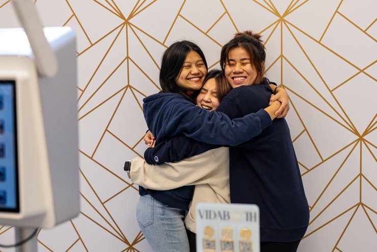 Three young women wearing casual clothes smile with their eyes closed as they hug each other in front of a geometric.