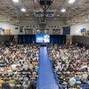 A large audience seated in a gymnasium faces a stage with a speaker and a screen displaying a presentation.