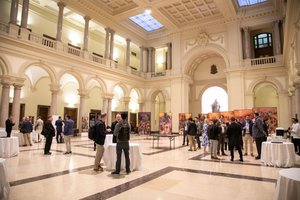 People are mingling and talking in a large, ornate hall with arched columns and paintings displayed along the walls.