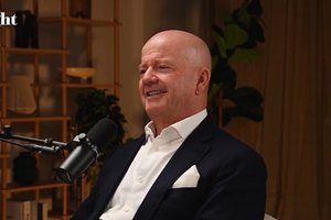 A bald man in a dark suit and white shirt smiles while speaking into a microphone in a warmly lit room.