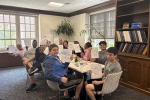 Ten smiling students sit around a table, proudly showing off their individual drawings of gingerbread people.