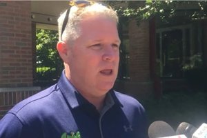 A man with short gray hair wearing a navy blue shirt speaks into microphones outside near a brick building and trees.