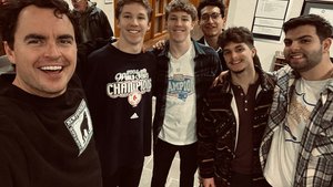 Six young men stand closely together indoors, smiling at the camera as one takes a selfie.