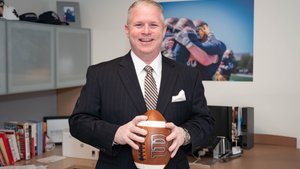 A man in a dark pinstripe suit smiles while holding a football in an office with sports photos and books.