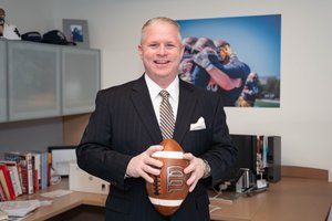 A man in a dark pinstripe suit smiles while holding a football in an office with sports photos and books.