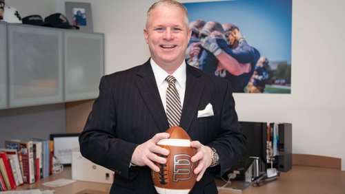 A man in a dark pinstripe suit smiles while holding a football in an office with sports photos and books.