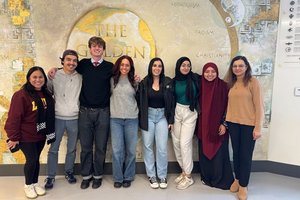 Eight people stand side by side smiling in front of a wall mural featuring a globe and religious symbols.