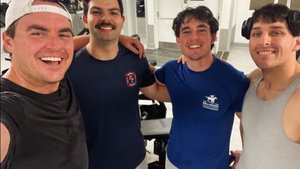 Four young men smile with their arms around each other inside a gym with workout equipment and mirrors behind them.