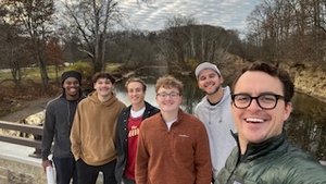 Six young adults stand close together on a wooden bridge over a calm creek, smiling at the camera on a cloudy day.