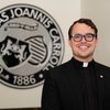 A man wearing glasses and a black clerical collar smiles in front of a circular university emblem on a beige wall.