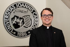 A man wearing glasses and a black clerical collar smiles in front of a circular university emblem on a beige wall.