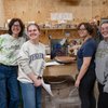 Four people smile and pose in a workshop with shelves of containers and a wooden table holding pottery and art supplies.