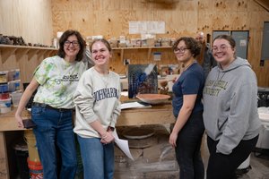 Four people smile and pose in a workshop with shelves of containers and a wooden table holding pottery and art supplies.