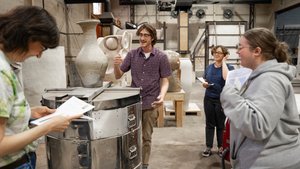 Four people stand around a large kiln in a workshop filled with ceramic sculptures, holding papers and smiling.
