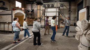 A group of six people stand inside a workshop with kilns and wood stacks while one person gestures and speaks to the others.