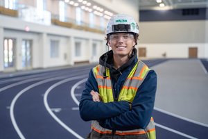 A young man wearing a white hard hat, safety glasses, and a reflective vest stands with arms crossed on an indoor running.