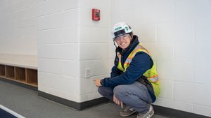 A person wearing a white hard hat, safety glasses, and a yellow safety vest is crouching and pointing at an electrical.