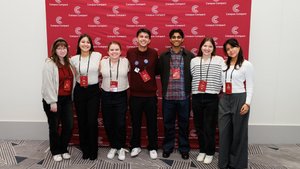 Seven young adults stand side by side smiling in front of a red Campus Compact backdrop indoors.