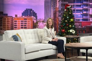 A woman in a white blazer and dark pants sits on a light sofa in a studio with a decorated Christmas tree behind her.