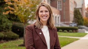 A woman with shoulder-length blonde hair smiles at the camera while standing outside near a tree with autumn leaves.
