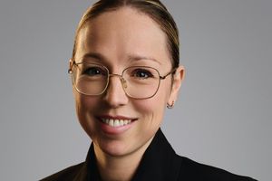 A woman with light skin and hair pulled back wears glasses, a black blazer, and a gold necklace while smiling.