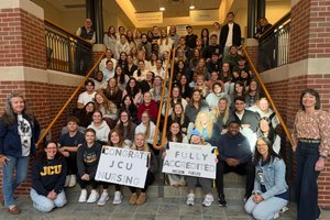 A large group of people sitting and standing on stairs inside a building hold signs that say Congrats JCU Nursing and Fully.