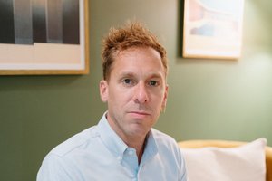 A man with short, tousled hair and a light blue button-up shirt sits on a yellow couch with his hands clasped.