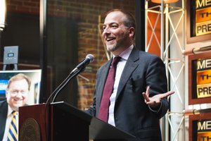 A man in a suit and tie smiles and gestures with one hand while speaking at a podium with a microphone.