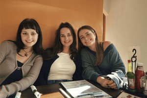 Three young women sit closely together at a wooden table in a restaurant, smiling at the camera.
