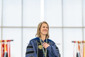 A university leader, wearing academic regalia and a ceremonial chain, smiles with her hand over her heart during
