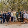 A group of young people stand outdoors on a paved area holding shovels and rakes with trees and playground equipment behind.