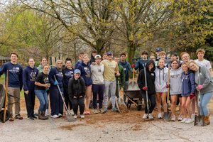 A group of young people stand outdoors on a paved area holding shovels and rakes with trees and playground equipment behind.