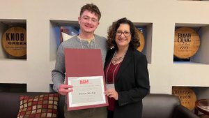 A young man and a woman stand smiling indoors, with the man holding a certificate and wooden barrels displayed behind them.