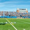 A football field with players on the sideline and spectators filling the blue stadium seats under a clear sky.