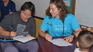 A woman in a blue John Carroll University shirt smiles while helping another woman fill out forms at a table as a child.