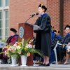 A woman in academic regalia speaks at a podium outdoors during a graduation ceremony with seated faculty behind her.