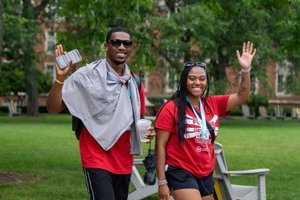 Two people wearing red shirts smile and wave while walking on a grassy area with trees and buildings behind them.