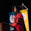 A speaker in red academic regalia, with an Alpha Sigma Nu stole, addresses an audience from a wooden podium at a university