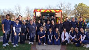A group of people in medical and emergency uniforms pose smiling in front of an open ambulance on a sunny day.