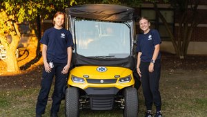 Two young women in navy blue JCU EMS shirts stand smiling beside a yellow emergency response vehicle outdoors.