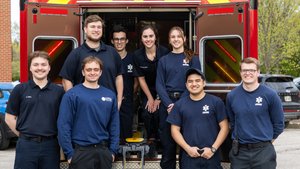 Eight young adults in navy blue uniforms smile while standing in and around the open back of a red emergency vehicle.