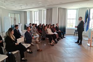 A man in a suit stands speaking to a seated audience of young adults in a bright room with large windows and flags.