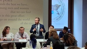 A man in a suit stands speaking into a microphone at a panel table while others listen and take notes in a conference room.