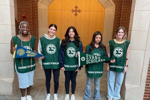 Five young women wearing green Evans Scholar vests stand in a row inside a brick and stone building holding college pennants.